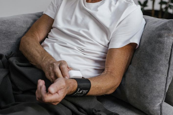 Senior adult monitoring blood pressure while sitting on a grey sofa indoors.
